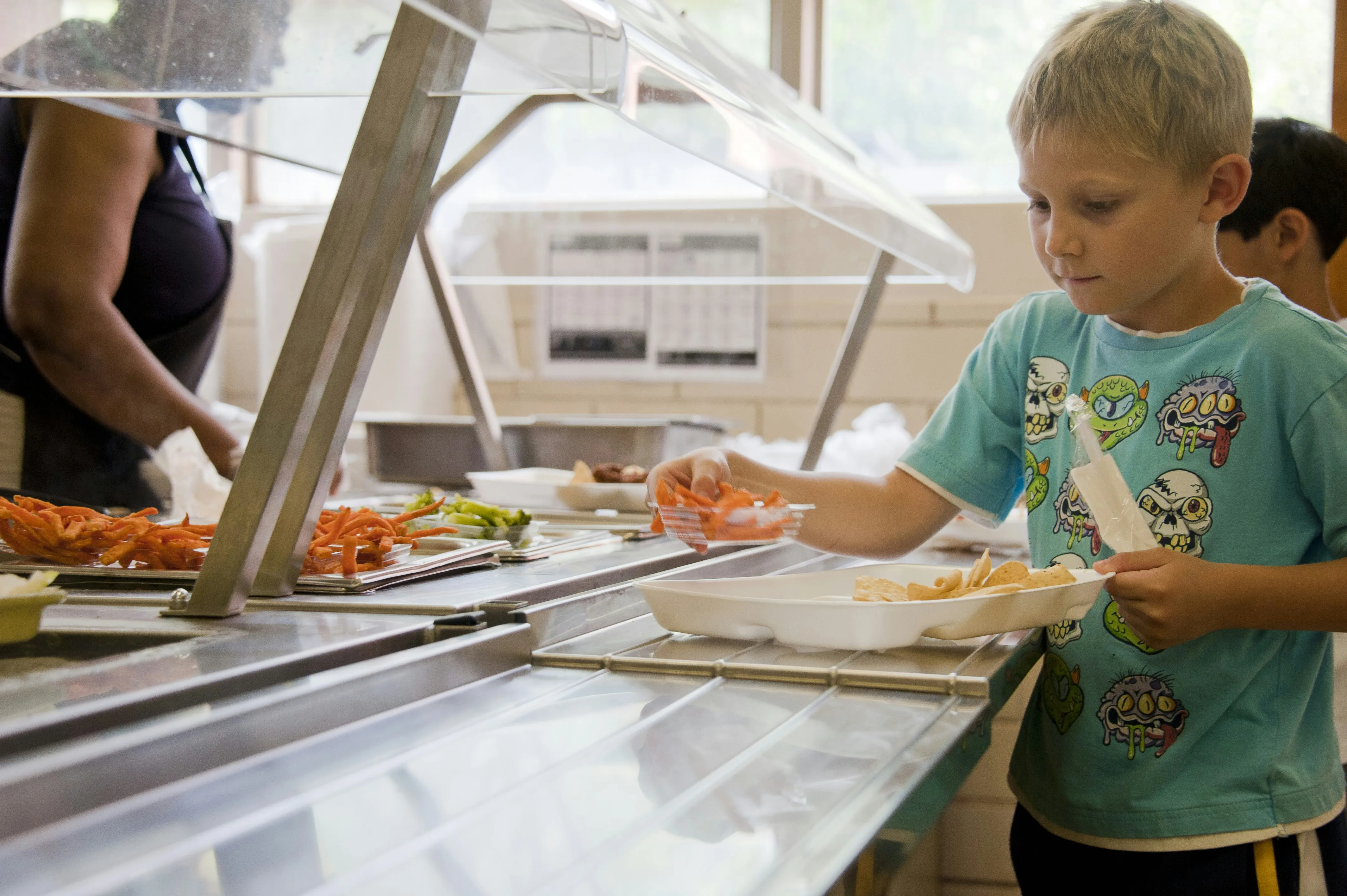 Young boy serving food at cafeteria cafeteria food counter with trays of vegetables and chips.