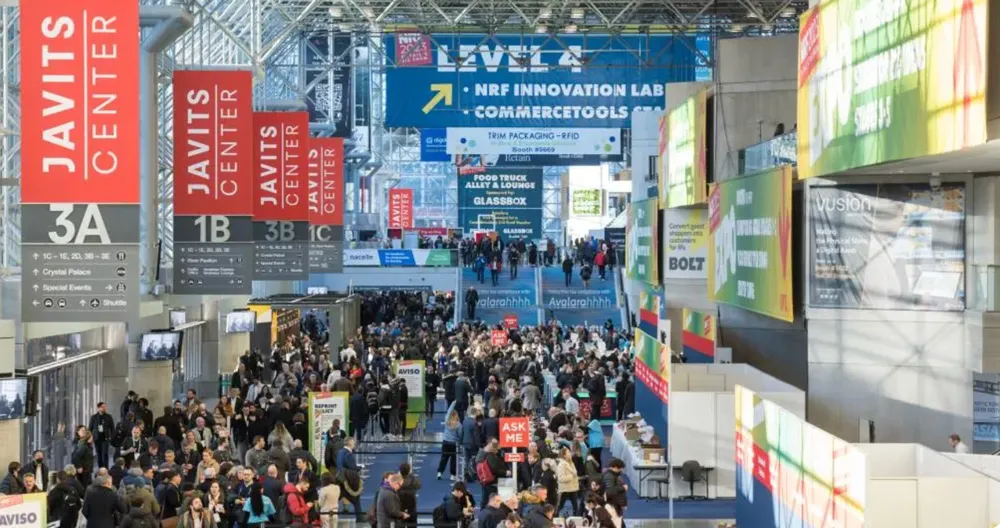 Crowded convention hall at Javits Center with hanging signs and many attendees walking and milling.