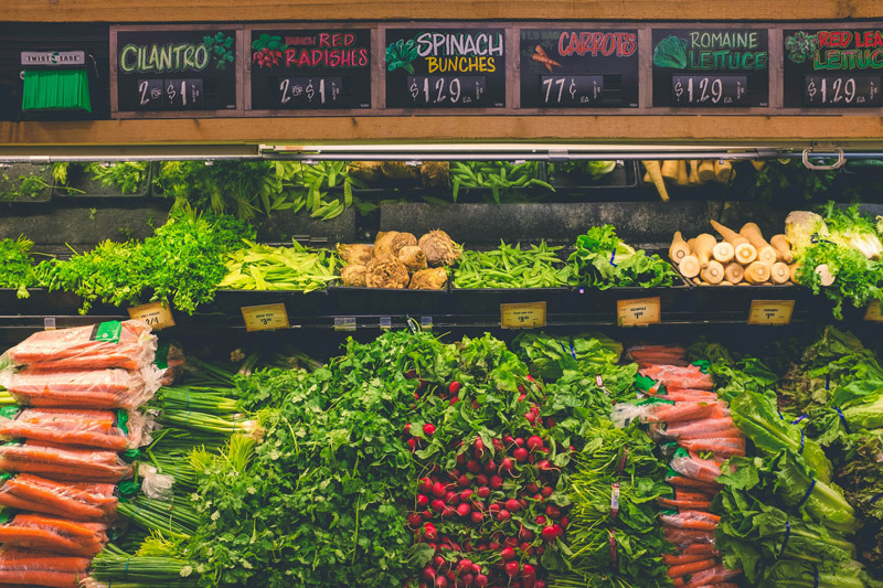 Grocery store produce section with fresh cilantro, radishes, spinach, carrots, and lettuce displayed with signs.