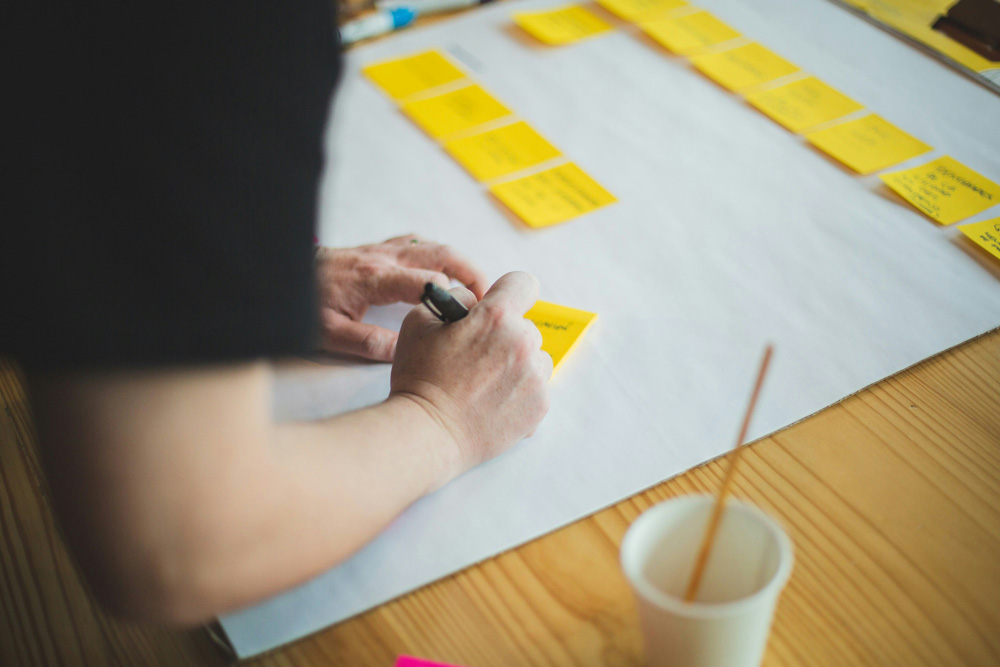 Person writing on a yellow sticky note at a table covered with organized sticky notes on white paper.